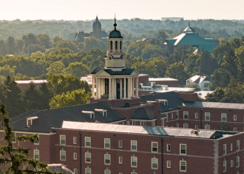 A brick building with a central cupola, surrounded by trees and additional buildings in the background.