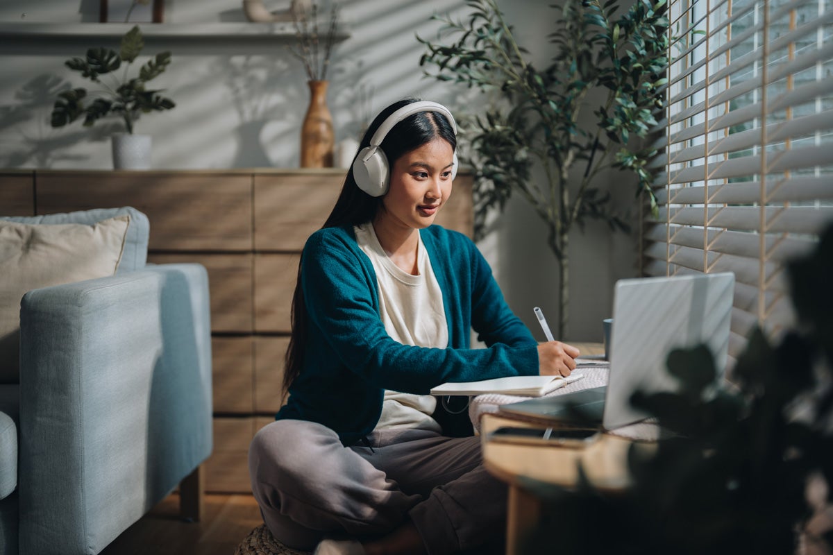 Woman wearing headphones, sitting on the floor, taking notes while watching a laptop in a cozy living room.