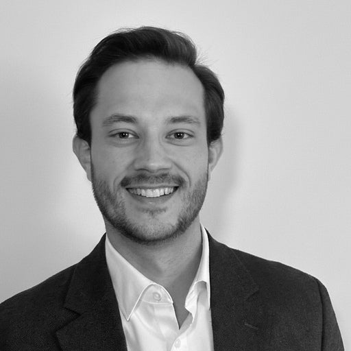 Smiling man with short dark hair and stubble, wearing a suit jacket and open-collared shirt, against plain background.