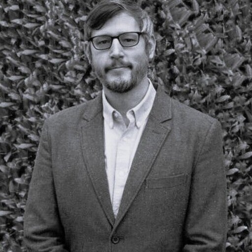 A man with glasses and a beard wearing a suit jacket stands in front of a leafy background.