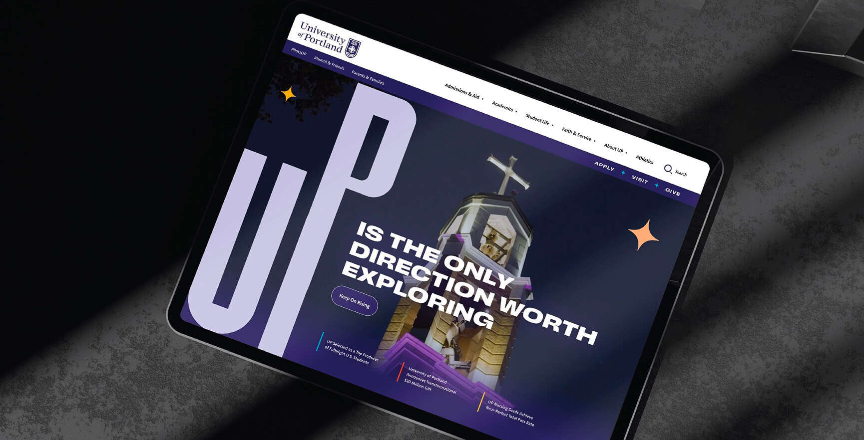 A tablet displays a university website with the text “UP IS THE ONLY DIRECTION WORTH EXPLORING.”.