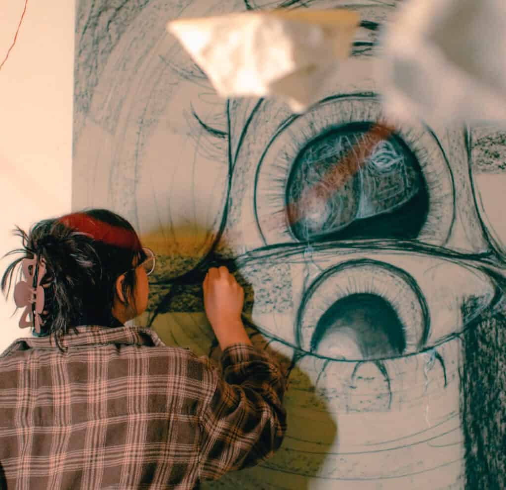 Person drawing large abstract eyes on a wall, wearing glasses and plaid shirt, with paper shapes hanging above.