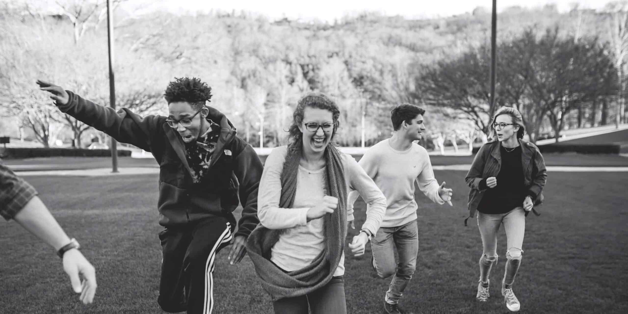 Four young adults run and laugh together outdoors on grass, with trees and hills in the background.