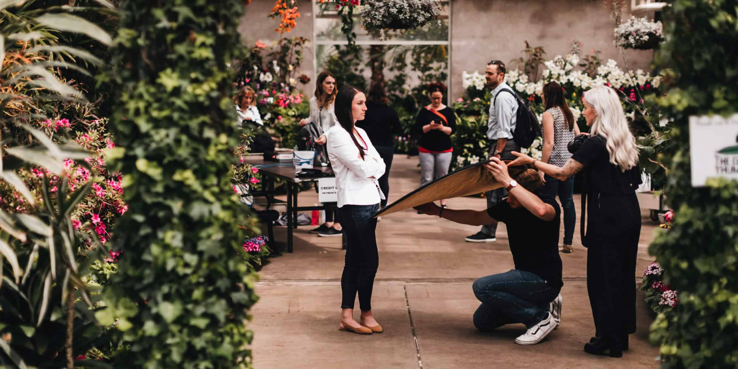 Several people gather in a greenhouse while a photographer adjusts equipment near a woman posing.