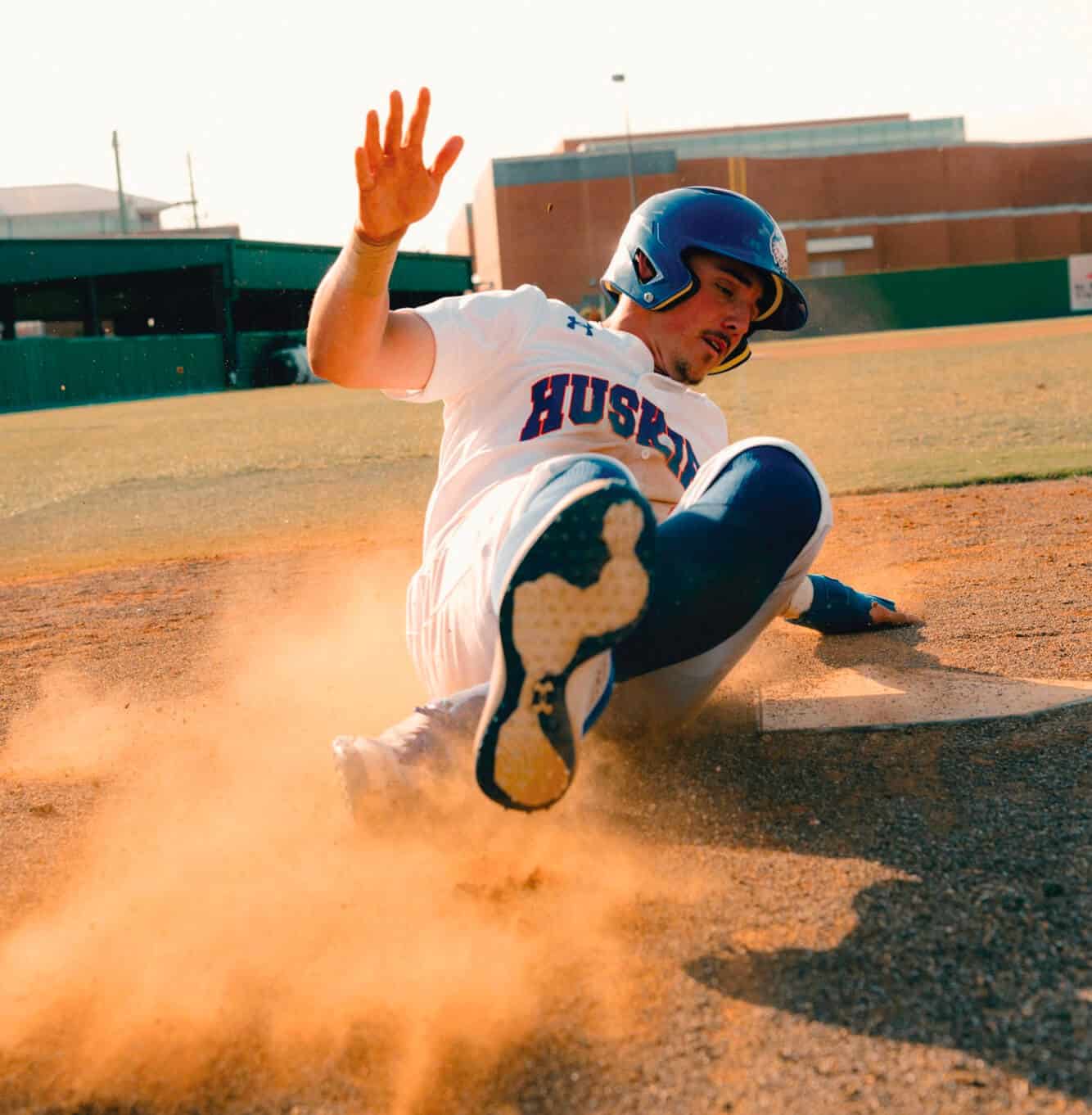 Baseball player sliding into base, kicking up dirt, wearing a helmet and "Huskies" uniform.