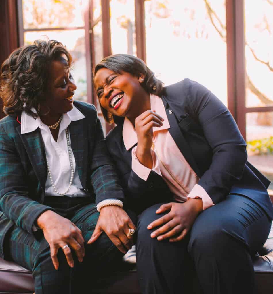 Two women in suits sit together, smiling and laughing in a brightly lit room with large windows.