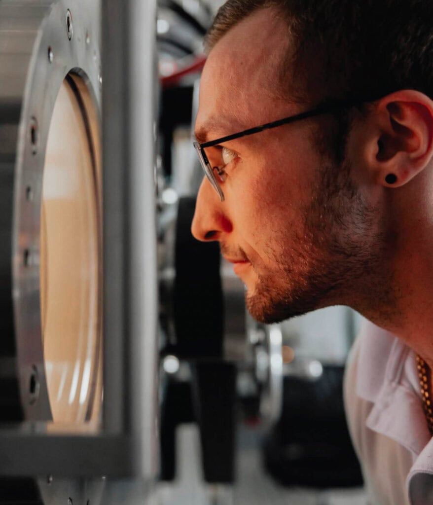 A man with glasses closely looks into a round window on a metallic machine, appearing focused.