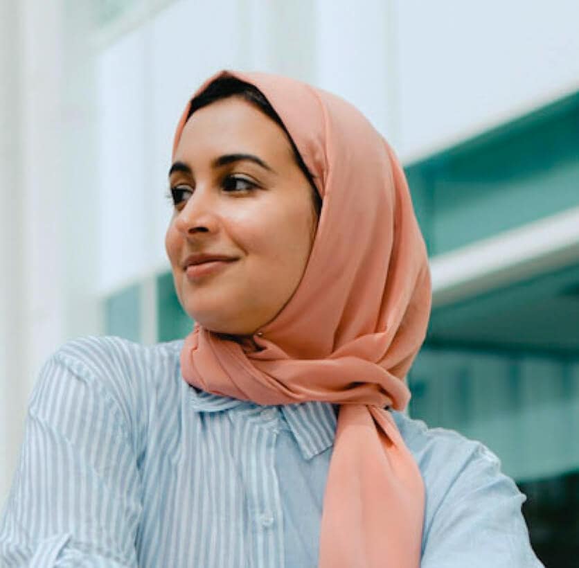 Woman in a pink hijab and striped shirt smiling and looking to the side in front of a modern building.