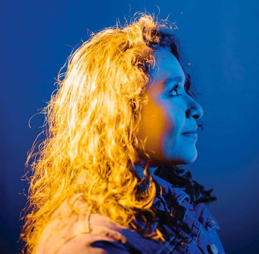 Woman with curly hair in dramatic blue and yellow lighting, looking upwards and smiling in profile.