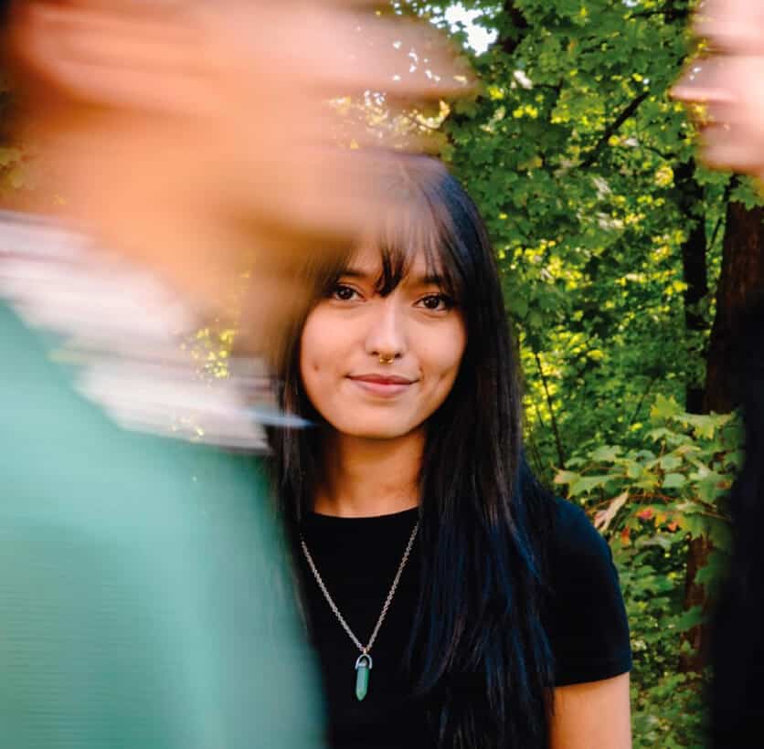Woman with long dark hair stands still, while blurred people move past her in a green, leafy outdoor setting.