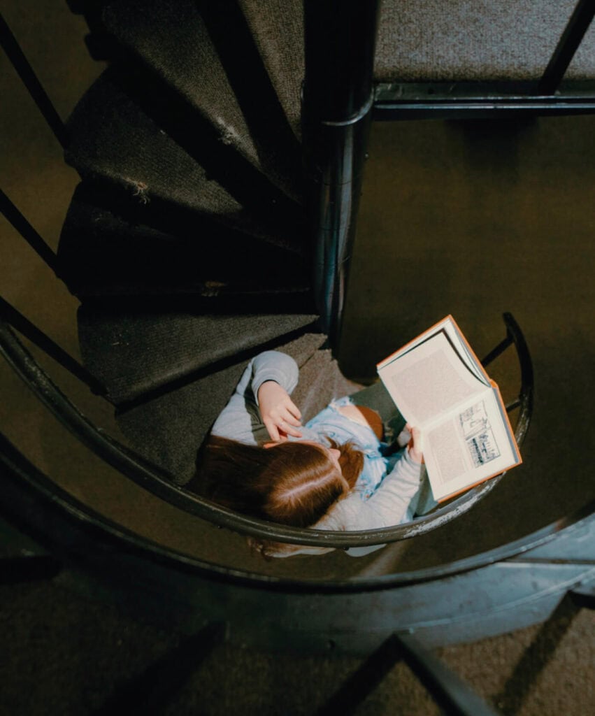 A person sits on a spiral staircase, reading a book, viewed from above.
