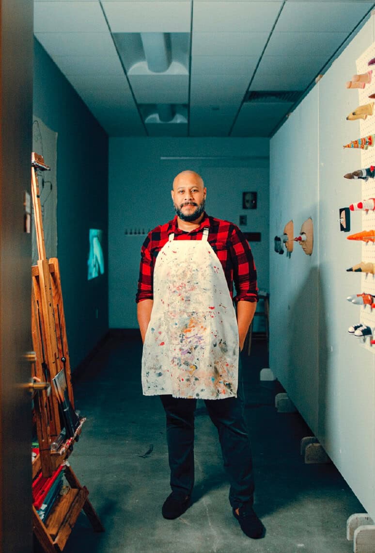 A man in a paint-splattered apron stands in an art studio, facing the camera with hands in his pockets.