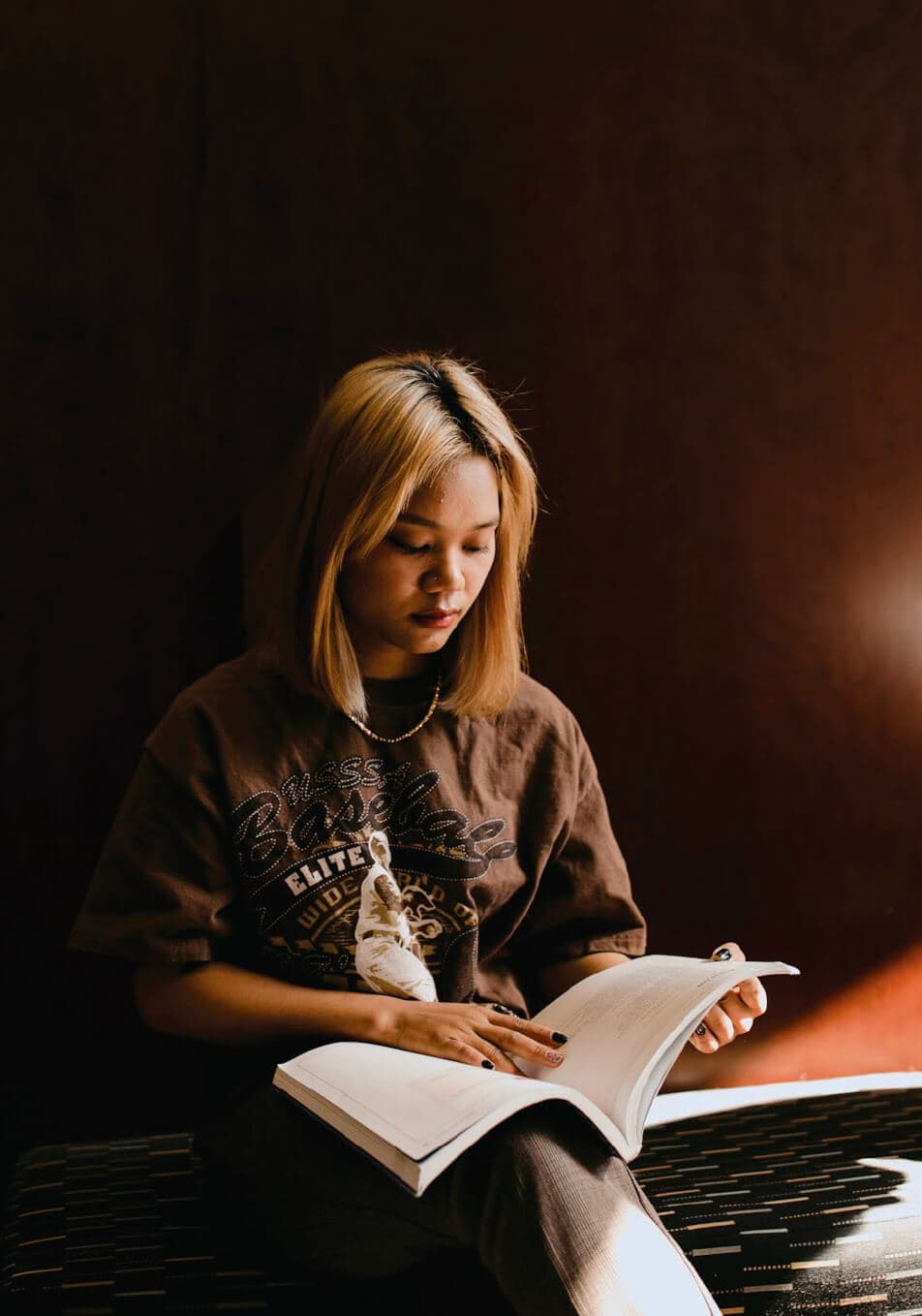 Young woman with blonde hair reading an open book in soft sunlight against a dark background.