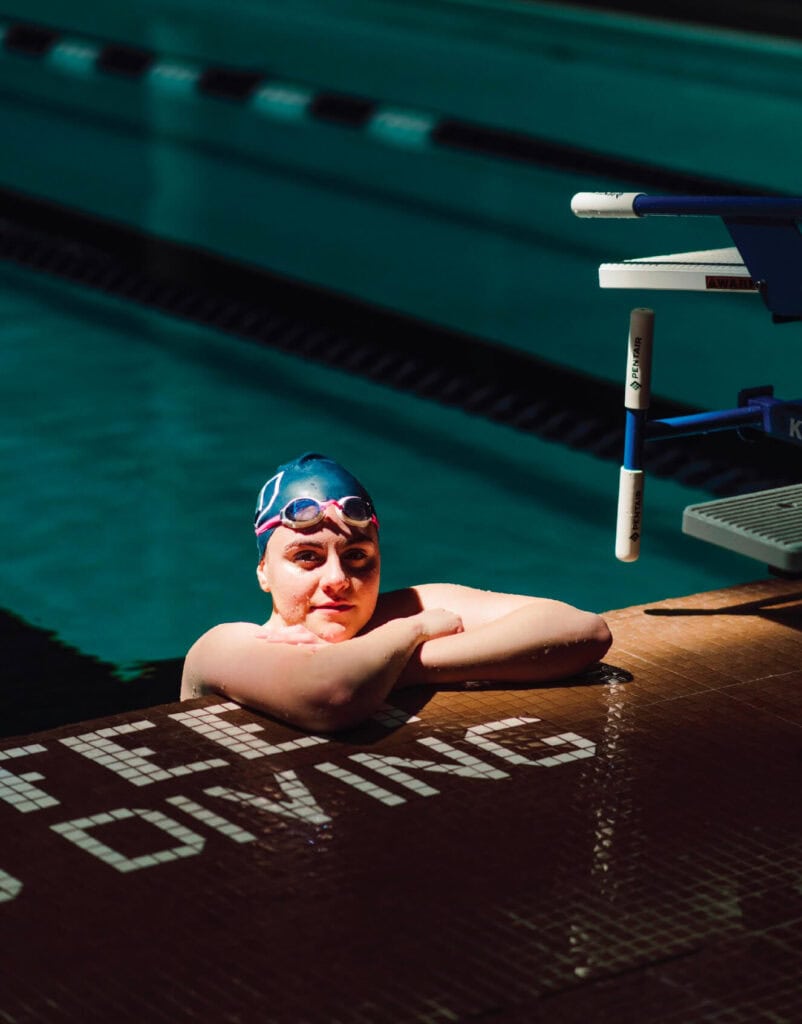 Swimmer in a blue cap and goggles rests at the edge of an indoor pool near a "No Diving" sign.