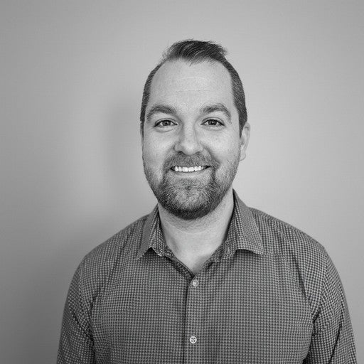 A smiling man with short hair and a beard, wearing a checked shirt, poses against a plain background.