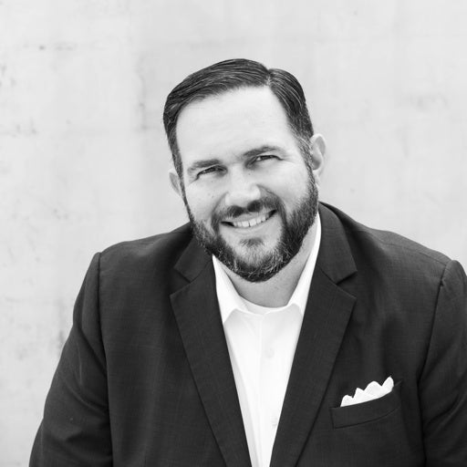 Man in a suit with a beard, smiling while sitting against a plain light-colored background.