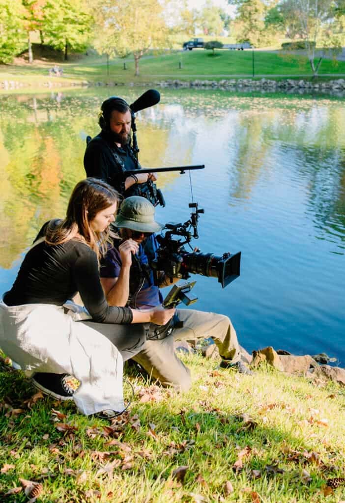 Three people filming with cameras and audio equipment by a pond on a sunny day.