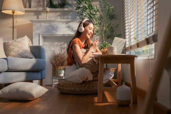 Young woman wearing headphones, sitting on the floor, smiling at a laptop in a cozy, sunlit room.