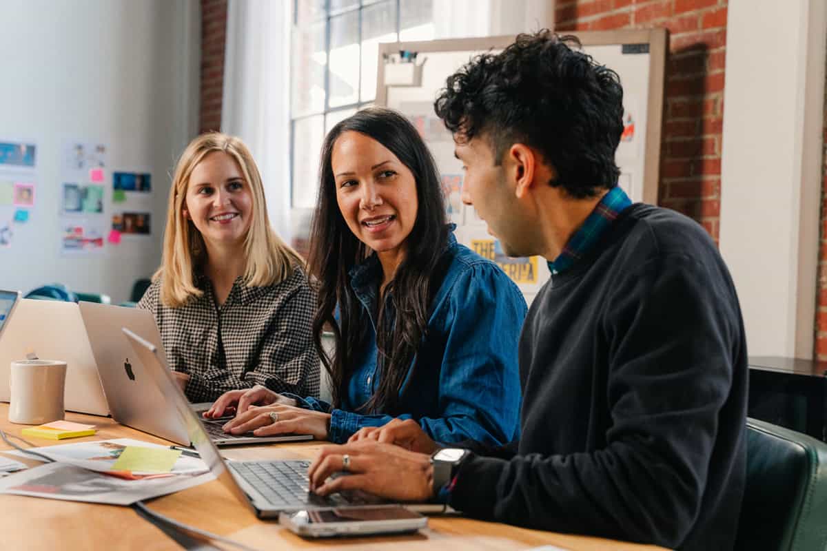 Three people sit at a table with laptops, talking and smiling in a brightly lit office space.