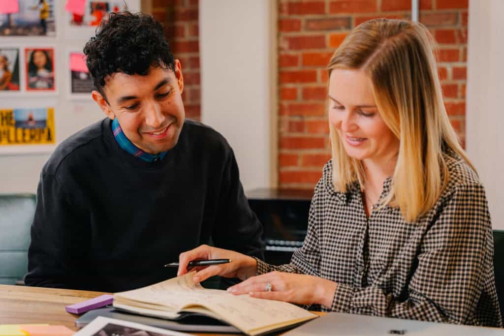 Two people sit at a table, smiling and discussing notes in an open notebook together.