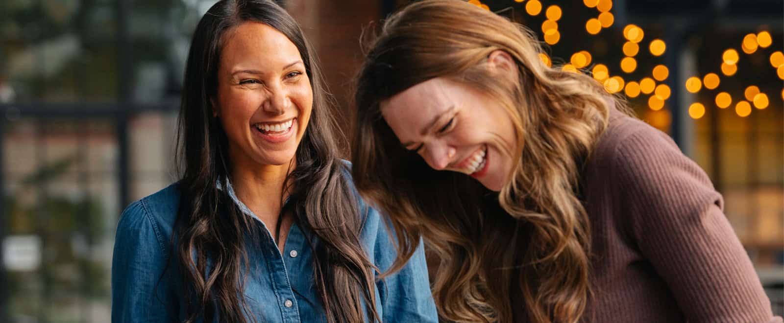 Two women laughing together outdoors under branded string lights, with the warm glow blurred in the background.