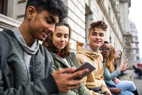 A group of teenagers sitting outside, smiling and looking at a phone together.