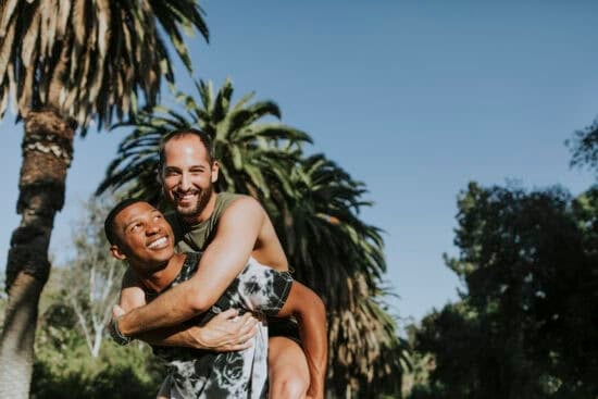 Two men smiling, one giving the other a piggyback ride outdoors with palm trees in the background.