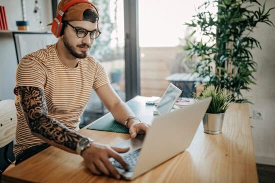Young man with headphones and a tattoo works on a laptop at a wooden desk in a modern, bright room.