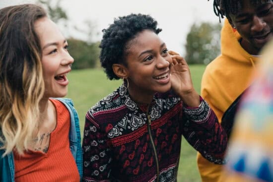 Three young people standing close together outside, smiling and laughing, with trees in the background.