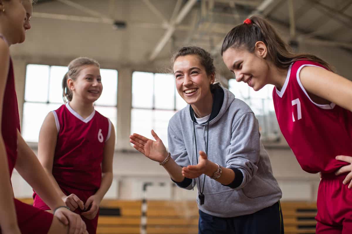 A coach enthusiastically talks to her young basketball team in red uniforms inside a gym.
