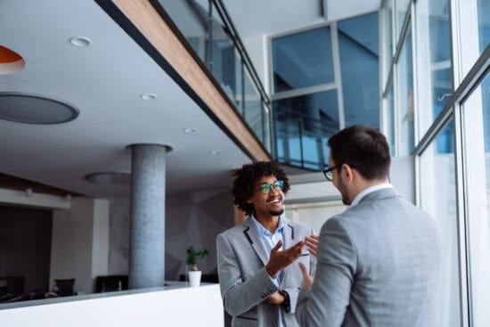 Two men in suits talking and smiling in a modern office with large windows and natural light.