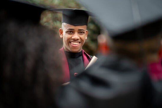 A smiling graduate in cap and gown holds a diploma, surrounded by other graduates.