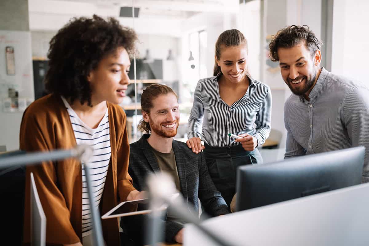 Four colleagues smiling and collaborating around a computer screen in a modern office setting.
