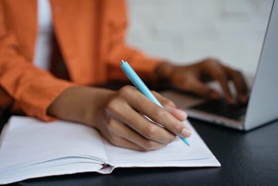 Person writing in a notebook with a pen while using a laptop, sitting at a desk.