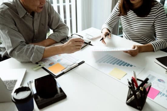 Two people sit at a desk reviewing charts and graphs, discussing business documents and data analysis.