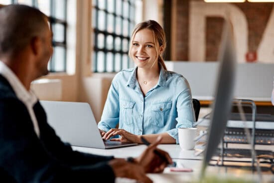 Woman smiling and typing on a laptop at a modern office desk, with a coworker in the foreground.