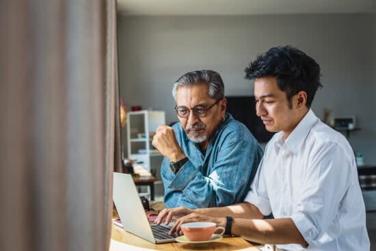 Older man and younger man sitting at a table, using a laptop together in a bright room.