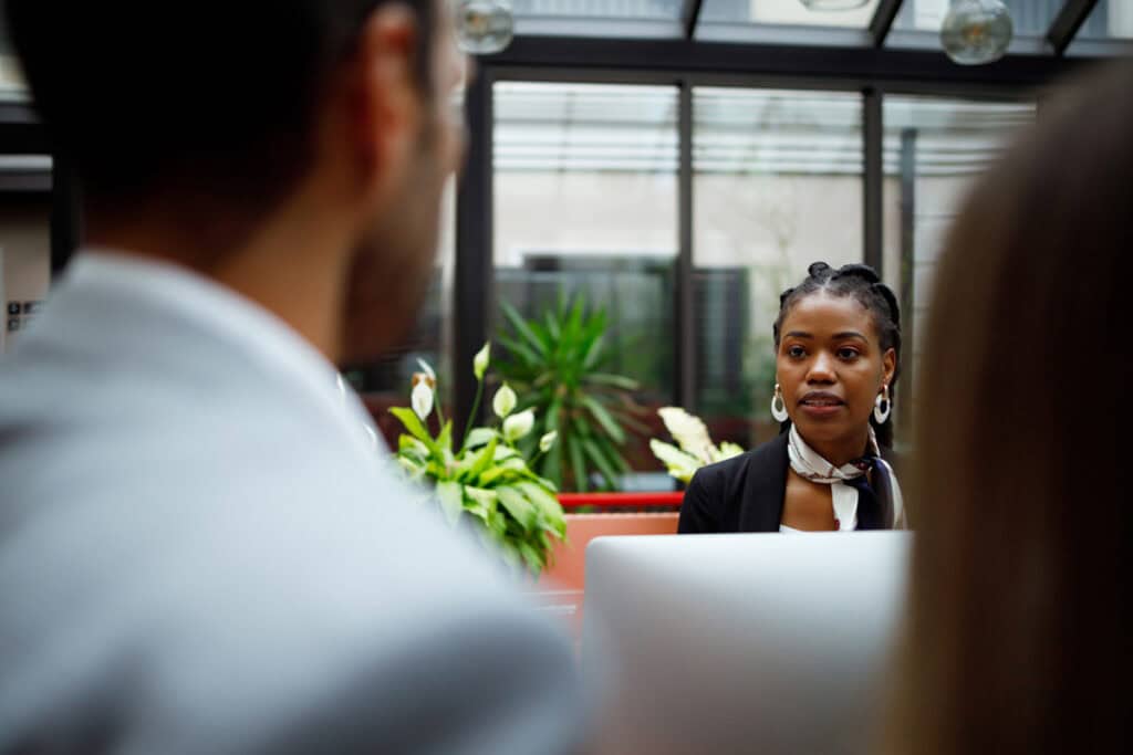 A woman in business attire listens attentively to colleagues in an office with plants in the background.