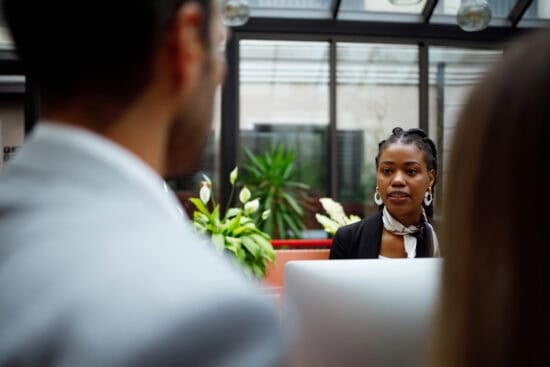 A woman in business attire listens attentively to colleagues in an office with plants in the background.