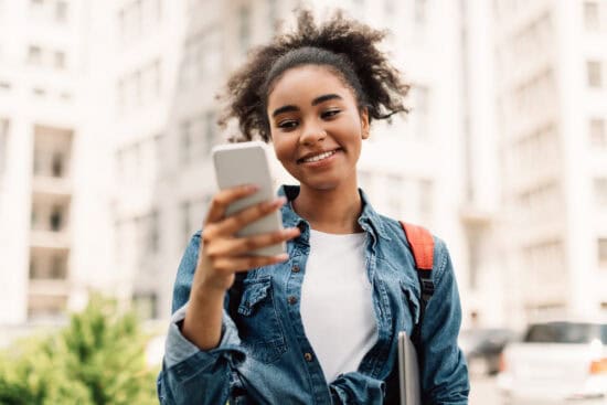 Smiling student outdoors looks at her phone, holding a laptop and wearing a backpack.
