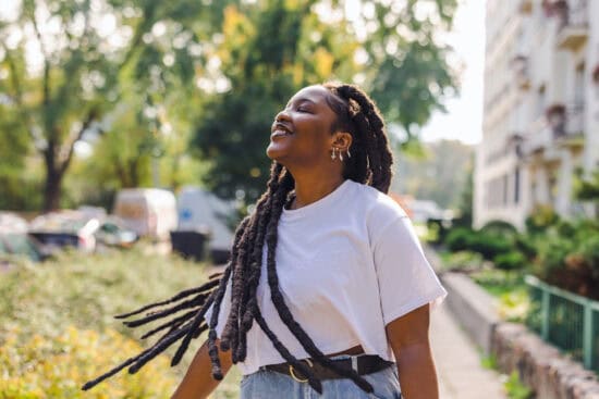 Smiling woman with long braids enjoys sunshine outdoors, wearing a white t-shirt and jeans.