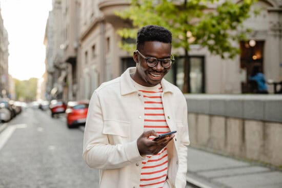 Man smiling while looking at his phone, walking on a city street lined with cars and buildings.