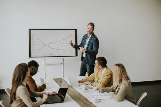 A man presents a graph to four colleagues seated at a table with laptops in a meeting room.
