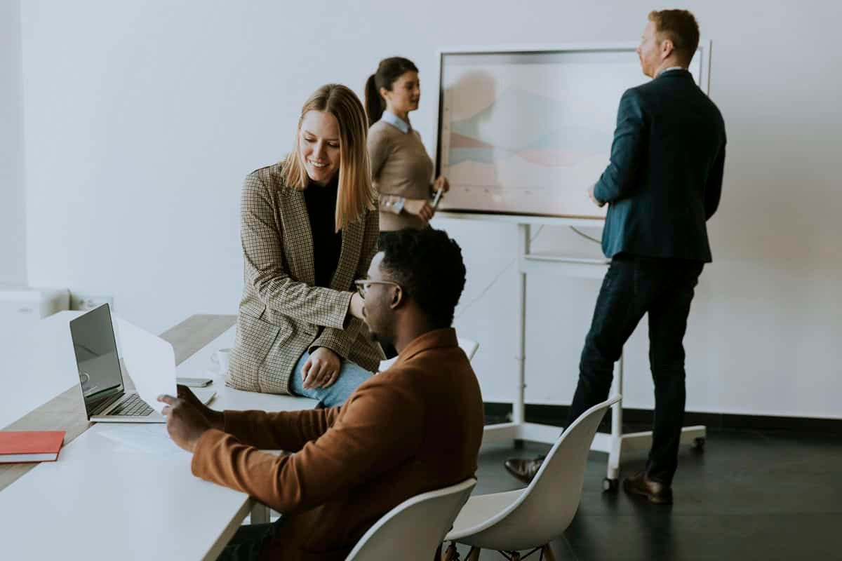 Four colleagues collaborate in an office, two at a desk with a laptop and two near a presentation board.