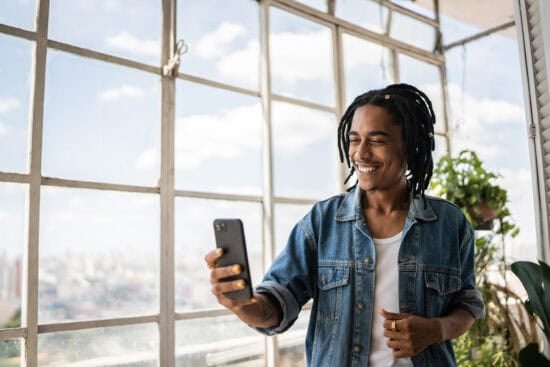Smiling young man with dreadlocks takes a selfie by large windows in a bright, urban indoor space.