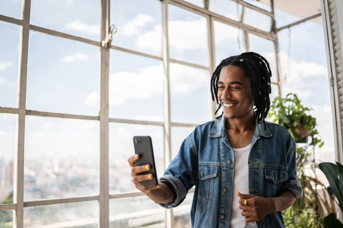 Smiling young man with dreadlocks takes a selfie by large windows in a bright, urban indoor space.