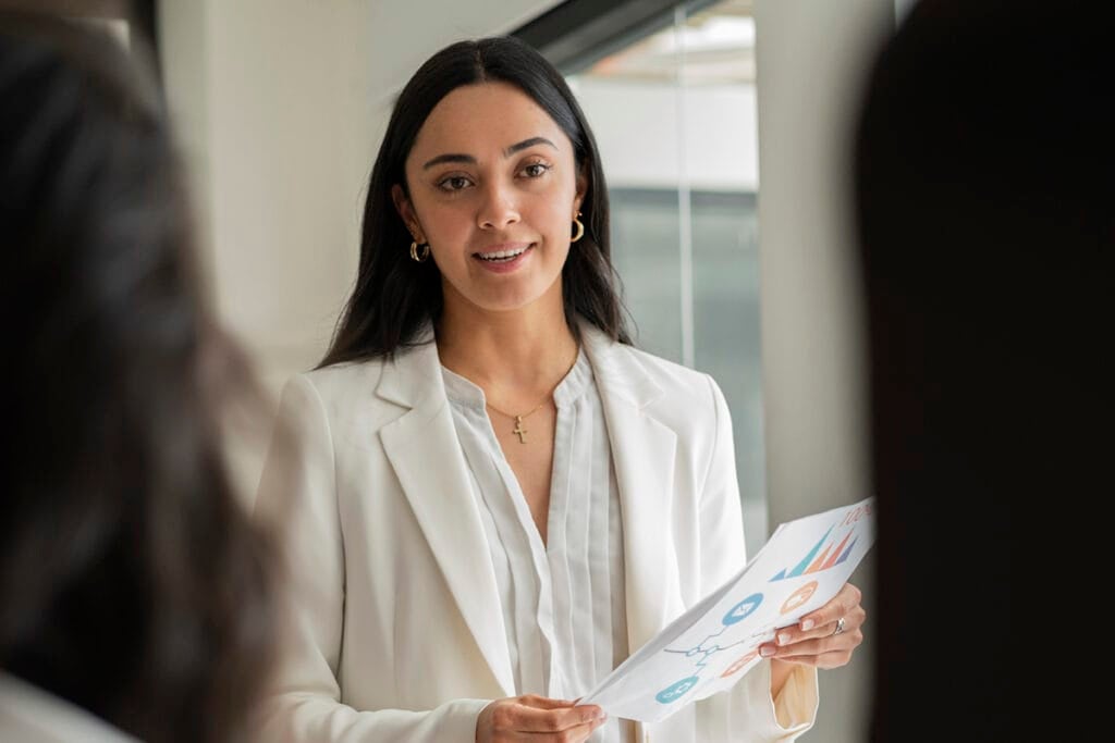 Woman in a white blazer holding papers, speaking in a bright office setting.