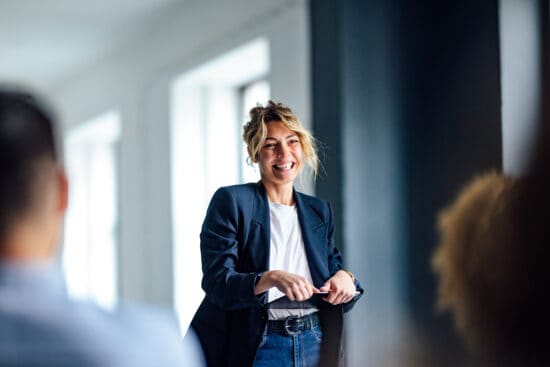 A woman in a blazer smiles and speaks in front of a group in a bright, modern room.