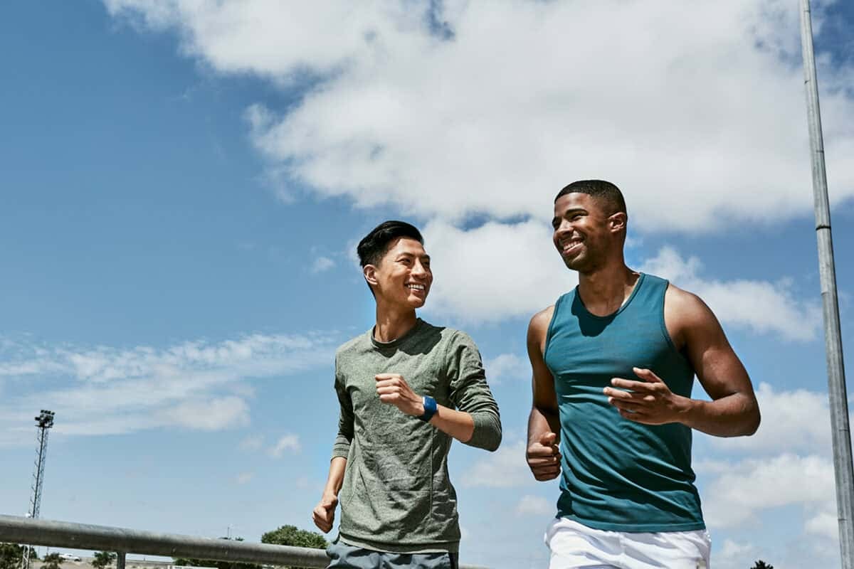 Two men jog outdoors on a sunny day, smiling and enjoying their run under a blue sky with clouds.