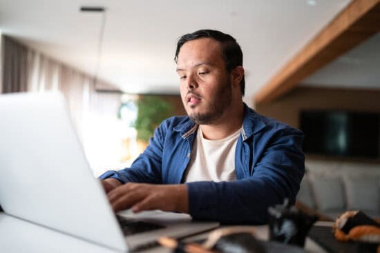 Man with Down syndrome using a laptop at a table in a bright, modern living room.